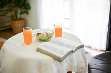 Close-up image of an opened book, a salad bowl, and two glasses of orange juice are on a table