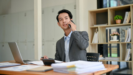 A smart millennial Asian businessman in a formal business suit is on the phone in his office.