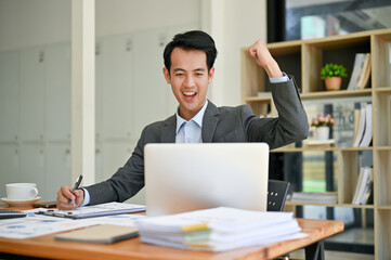 Fototapeta premium Cheerful businessman is raising his hand, looking at his laptop screen, and celebrating his success