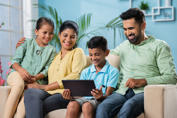 Happy Indian parents sitting with sibling kids while son using digital tablet at home - concept of family bonding or time and weekend holidays and cyberspace