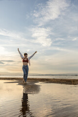 Obraz premium woman on the beach dancing at sunset in backlight with the sun behind in shadow, woman on vacation walking on the sand with golden water reflections from the sun at sunset