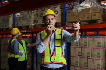 Group of warehouse workers with hardhats and reflective jackets using tablet, walkie talkie radio...