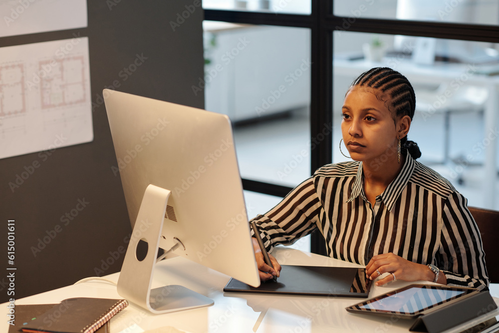 Aufkleber Young African American webdesigner or artist with stylus drawing sketch on touchpad screen while sitting by workplace in front of computer