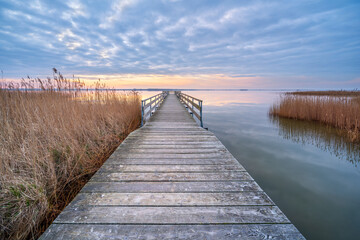 Calm Lake at Sunrise with Long Wooden Pier and Reeds under cloudy sky before sunrise