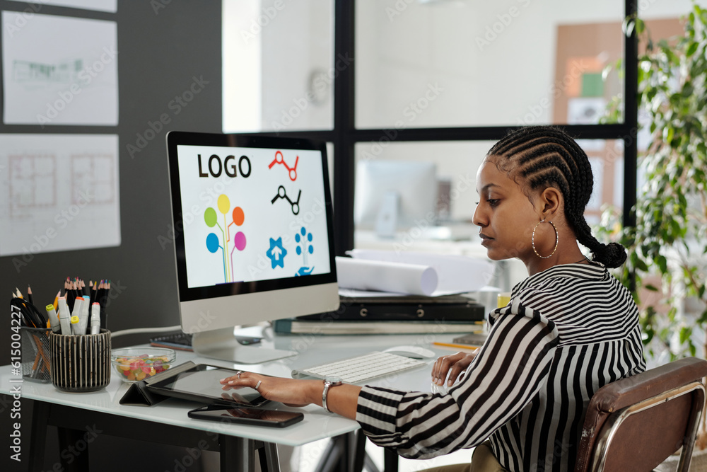 Poster side view of young serious african american female webdesigner sitting by workplace in front of comp