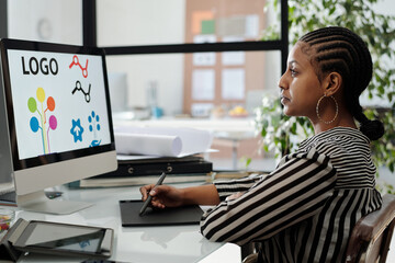 Serious African American female webdesigner sitting by desk in front of computer monitor with logo...