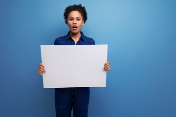 young pretty hispanic business woman with fluffy hair dressed in blue denim suit holding billboard with blank space