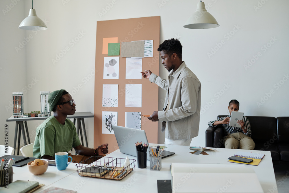 Poster young confident architect in casualwear pointing at noticeboard with sketches on papers while talkin