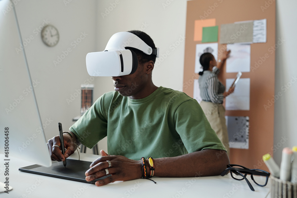 Poster Young African American male designer with vr headset using touchpad and stylus while sitting by desk and creating new computer games