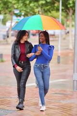 Two women, one African-American and one Latina smiling, walking together in the multicolored rain through a city park 
