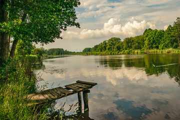 Widok na stary drewniany pomost na jeziorze, chmurki odbijające się w wodzie, zieleń drzew