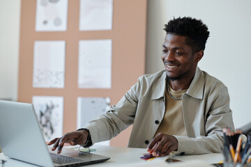 Young smiling African American male webdesigner pressing key of laptop keyboard while sitting by workplace and creating new website