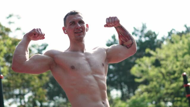 Low angle handheld shot of confident muscular sportsman with tattooed arm showing biceps while listening to music on earbuds during training on sports ground