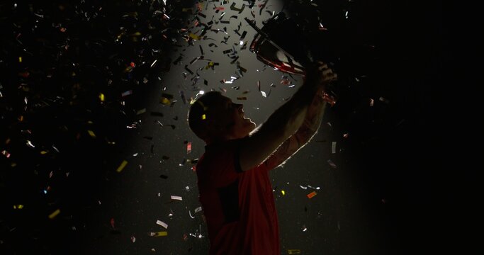 Silhouette of Caucasian male rugby player raising a trophy above head against bright light and falling confetti. Super slow motion, shot on RED cinema camera