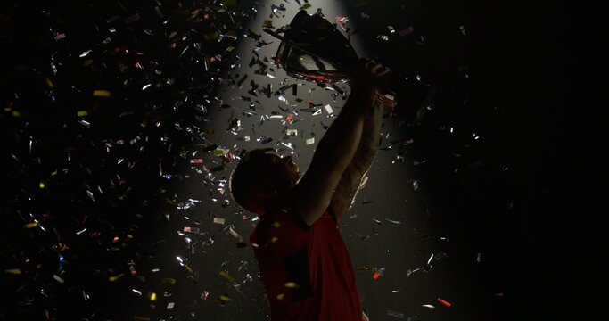 Silhouette of Caucasian male rugby player raising a trophy above head against bright light and falling confetti. Super slow motion, shot on RED cinema camera