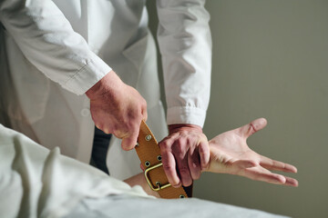 Close-up of male psychiatrist in lab coat binding hand of insane patient with belt to bed during...