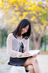 Portrait cute smiles Asian of attractive young teenage girl a holding diary book writing note at Yellow Tabebuia Chrysotricha flowers with the park in spring day at Evening background in Thailand.
