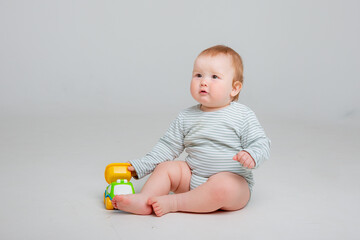 Cute toddler boy 10 months old in white bodysuit plays with car toy sitting on a white background, space for text
