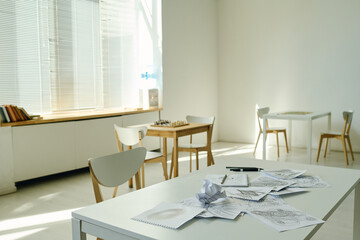 Part of spacious ward or room in mental hospital with chairs standing by desks with pencils and paper shets with drawings of sick patients