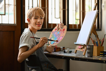 Young hipster queer artist holding palette and brush sitting in front of easel in bright studio. Leisure activity and art concept