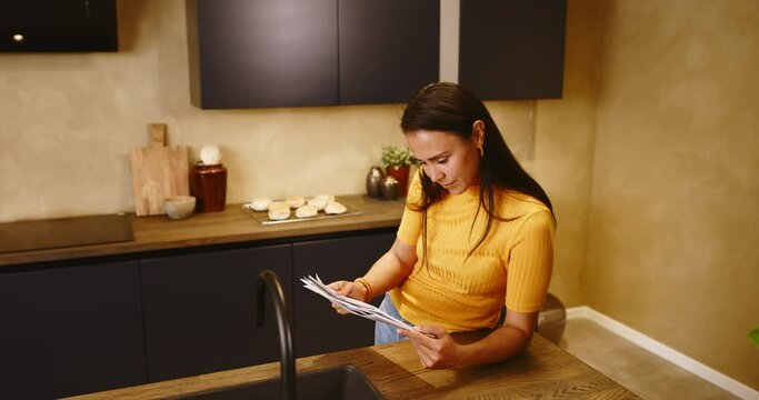 Young woman standing in her kitchen at home reading the financial pages of a newspaper