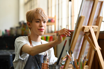 Satisfied young gay man painting on canvas in art workshop. Art, creative hobby and leisure...