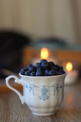 Cup of blueberries, set of books, phone, eyeglasses, lit candles and blanket on the table. Hygge at home. Selective focus.