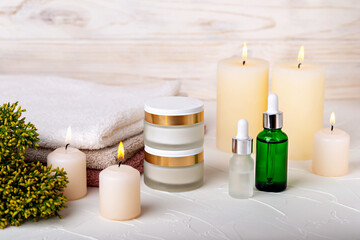 Obraz na płótnie Canvas Cosmetic jars with cream and dropper bottles among burning candles. Towels and plant complete spa concept. Care and relaxation, beauty and health. Selective focus