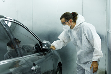 Young professional car painter wearing protective suit standing next to car in car painting room inspecting car body to look for scratches and damage