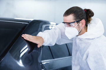 Fototapeta na wymiar Young professional car painter wearing protective suit standing next to car in car painting room inspecting car body to look for scratches and damage