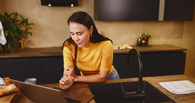 Smiling young woman leaning on a kitchen island at home and working online with a laptop