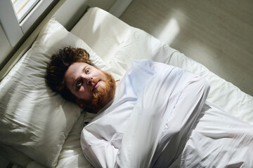 Above angle of young bearded man in straitjacket suffering from schizophrenia lying on bed in ward...