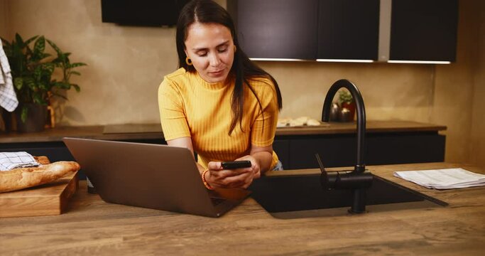 Young woman checking her phone while working online with a laptop at her kitchen island at home