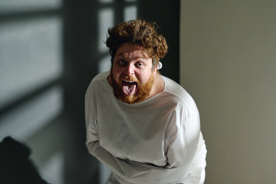 Young insane man in straitjacket showing tongue and screaming while standing in front of camera in mental hospital ward and looking at you