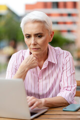 Obraz premium Pensive elderly woman working at computer, sitting at workplace, online work on street. Attractive freelancer working by workplace. Successful business concept
