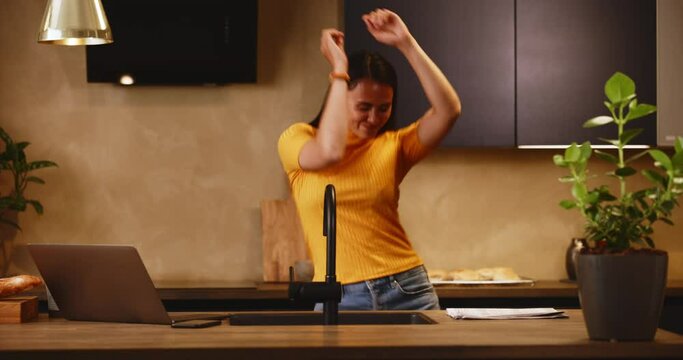 Smiling young woman working on a laptop and dancing in her kitchen at home