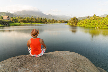 typed man with hat meditating sitting on a rock at the water's edge with panoramic view of the mountains in sierra nevada de santa marta colombia - obrazy, fototapety, plakaty