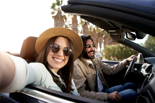 Caucasian young couple driving a cabrio car. The woman takes a selfie with palm trees in the background. They smile enjoying their vacation. She looks at the camera.