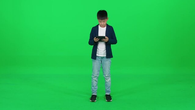 Focused asian schoolboy in a suit standing on a green background and using a screen tablet, reads an e-book, template on a chromakey.