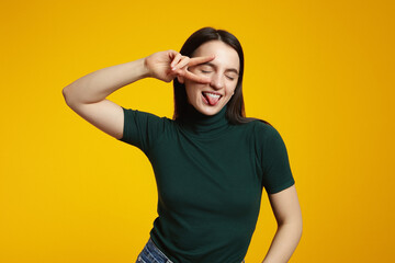 Portrait of a happy young girl showing peace gesture with closed eyes while sticking her tongue out isolated over yellow background