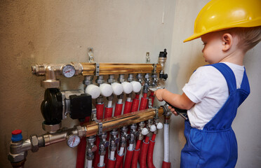 Baby boy in safety construction helmet using wrench tool while fixing plumbing installation with pipes and thermometer gauge. Kid plumber repairing plumbing pipe system in apartment under renovation.
