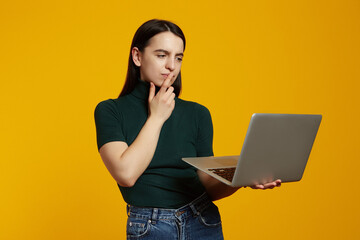 Portrait of a doubtful young girl holding laptop computer isolated over yellow background.