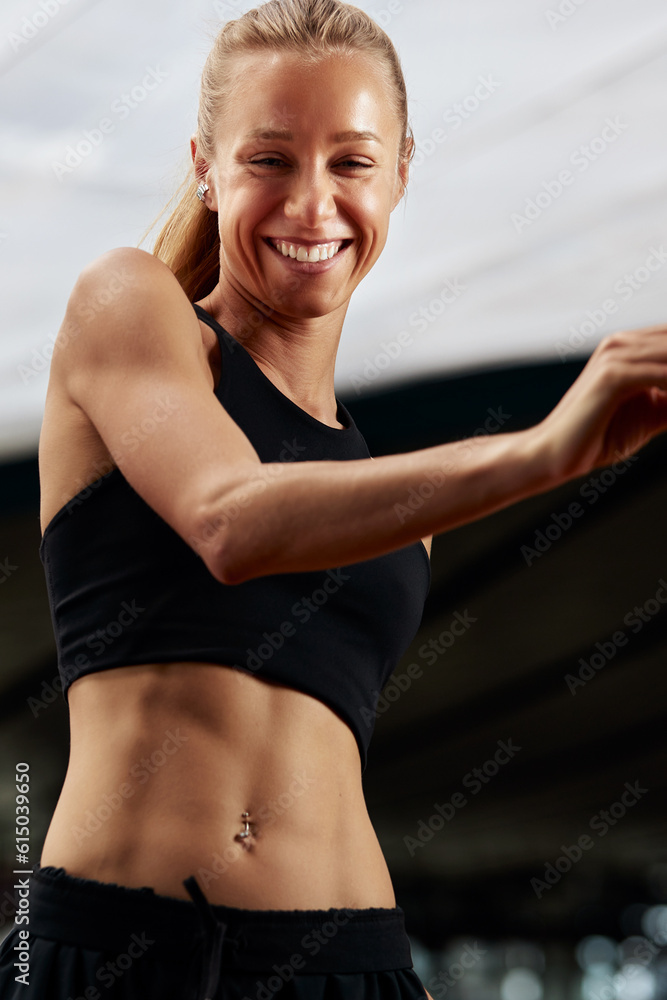 Wall mural Young fitness girl is warming up in the stadium. Attractive slender smiling blonde in a black tracksuit makes active movements with her arms and legs. Active lifestyle.