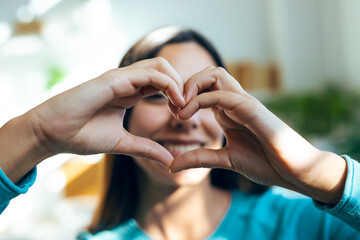 Romantic happy woman making heart shape with hands while smiling to camera at home