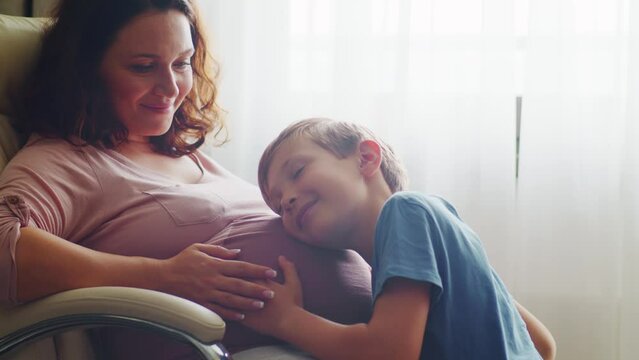 Child hugs and kisses her mother's pregnant belly. Happy son strokes the big belly his pregnant mom near window at home. Happy family life in an expectation a newborn. Pregnancy and maternity.