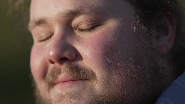 Young man closing eyes in meditation close up face sitting at park outdoors. Contemplative male caucasian overweight person feeling free from stress and worry