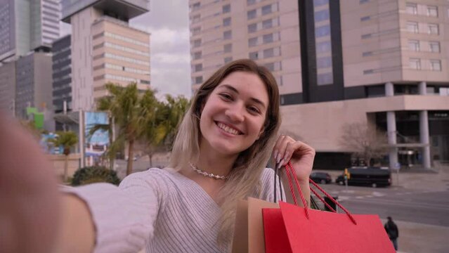 Young caucasian happy girl holding bag is taking a selfie to shy are in social media. Attractive blondie has been shopping during sales looking at camera with buildings of the city in the background