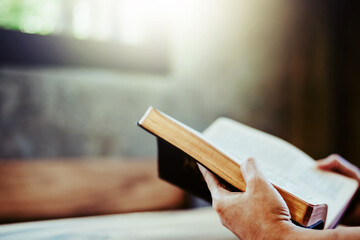 Close up of a human hands hold  while reading th open holy bible on wooden table with morning sunlight from the window, Christian devotional, faith and bible study concept with copy space
