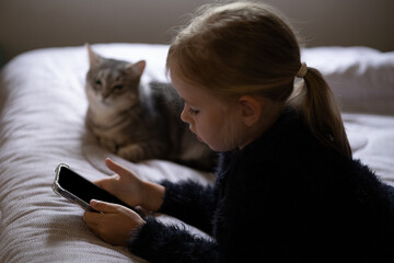 Little girl with blond hair with ponytail looking at smartphone with blank screen. Child and cat...