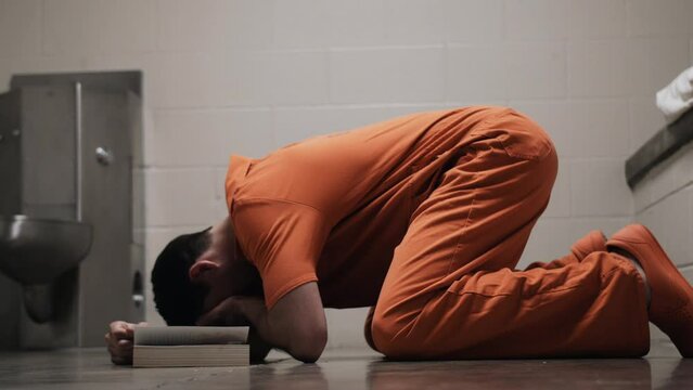 A sad prison inmate, man in prison wearing orange prison jumpsuit uniform on his knees crying with bible, praying, religious, God, christianity.